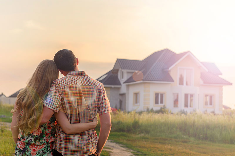 A couple stands outdoors with their arms around each other, looking at a modern house in the distance under a bright, hazy sky.