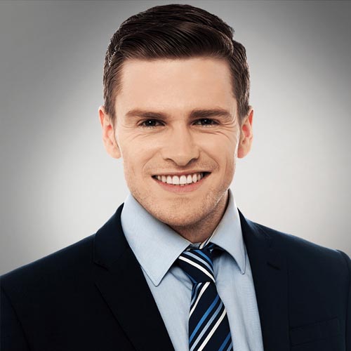 A young man in a dark suit, light blue shirt, and striped tie smiles confidently at the camera against a plain gray background.
