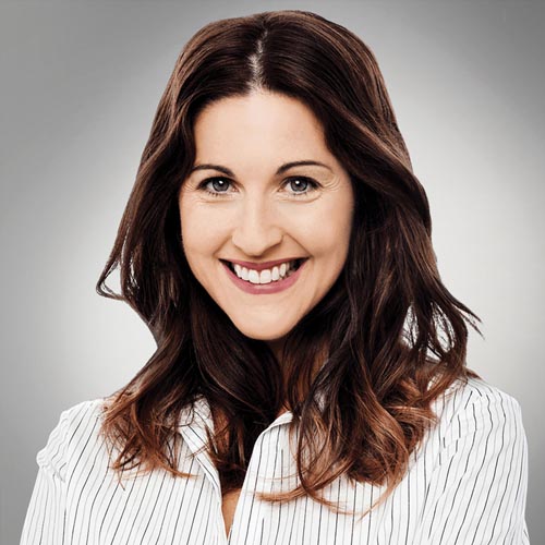 Smiling woman with long brown hair wearing a white shirt with thin black stripes, posed in front of a neutral gray background.